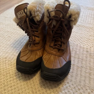 Bruna vinterboots från UGG med fårskinn - Säljer ett par bruna vinterboots från UGG med snörning, svart gummisula och mjukt fårskinnsfoder. Skorna är i skinn och har en robust, räfflad sula för bra grepp. Perfekta för kalla dagar och riktigt snygga till vinterlooken.