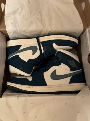 Nike Air Jordan 1 Mid blå/vit strl 42 - Nike Air Jordan 1 Mid sneakers i marinblått och vitt med klassisk Jordan-logga på sidan. Skorna har snörning, rund tå och platt sula. Tillverkade i skinn med perforerad tåbox för extra ventilation. Snygg kontrast mellan de blå och vita panelerna.