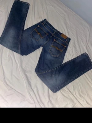 Blå jeans från Nudie Jeans Co - Säljer ett par klassiska blå jeans från Nudie Jeans Co med snygga kontrastsömmar och broderad logga på bakfickan. Modellen har rak passform och normal midja. Materialet är slitstarkt jeans och färgen är mörkblå med ljusare tvättade partier.