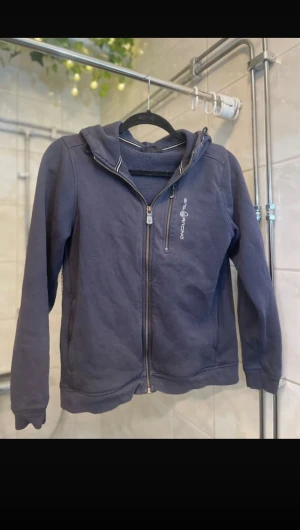 Marinblå hoodie från Sail Racing - Säljer en marinblå hoodie från Sail Racing med dragkedja framtill och en extra ficka med zip på bröstet. Tröjan har huva, långa ärmar och är gjord i mjuk bomull. Perfekt för chill dagar och har en clean look med diskret logga på bröstet.
