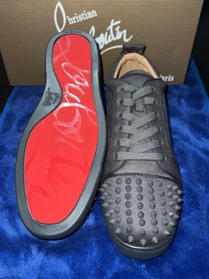 Christian Louboutin svarta sneakers med nitar - Säljer ett par svarta sneakers från Christian Louboutin med ikonisk röd sula och coola nitar på tån. Skorna har snörning och är tillverkade i mocka med detaljer i skinn. Perfekt för dig som vill sticka ut med en lyxig och edgy stil.
