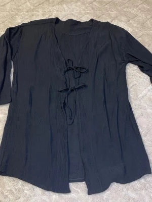 Svart kofta med knytband - Svart, tunn och luftig kofta med lång ärm och två knytband framtill. Materialet är lätt skrynklat och känns mjukt mot huden, perfekt att slänga över en t-shirt eller linne. Enkel och stilren design som funkar till många olika outfits.