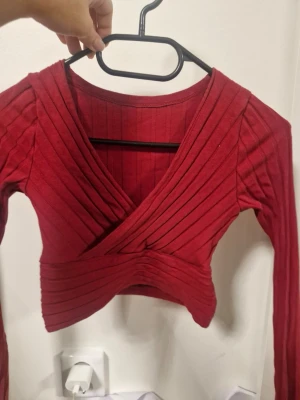 Röd omlott croppad topp - Säljer en röd croppad topp med omlott-design och ribbad struktur. Toppen har långa ärmar och djup v-ringning framtill. Materialet känns mjukt och stretchigt, perfekt för att styla med höga jeans eller kjol. Snyggt plagg för dig som gillar trendiga detaljer.