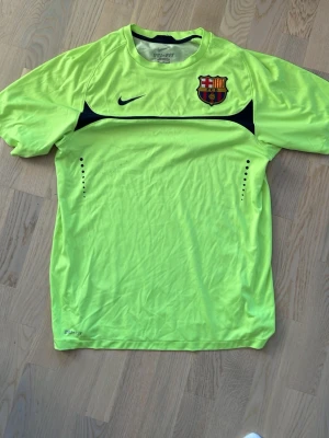 Barça Nike Dri-Fit träningströja M Retro säsong 2010/2011 - Snygg Barcelona träningströja från Nike i Dri-Fit material, perfekt för fotbollsträning. Tröjan är neon-gul med svarta detaljer och har klubbens emblem broderat på bröstet. Ventilationshål på sidorna och rund hals för extra komfort.