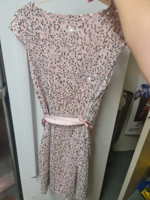 Rosa paljettklänning med rosett - Säljer en ljusrosa kortklänning från H&M, täckt med glittriga paljetter och ett matchande satinband som knyts i midjan. Klänningen har en rund halsringning och är ärmlös, perfekt för att sticka ut med extra bling.