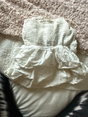 Vit bandeau tubtopp från Loavies - Supersöt vit bandeau tubtopp från Loavies i storlek S. Toppen har smockad överdel och volangdetaljer med broderade hålmönster. Perfekt för sommarens alla tillfällen, med en romantisk och fräsch vibe. Materialet känns lätt och luftigt.