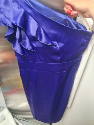 Blå oneshoulder klänning från Amisu - Säljer en snygg blå kortklänning från Amisu i storlek 38. Klänningen har oneshoulder-design med volangdetaljer längs axeln och är tillverkad i ett glansigt polyestermaterial som ger en lyxig känsla. Perfekt för dig som vill sticka ut med en cool och modern look.