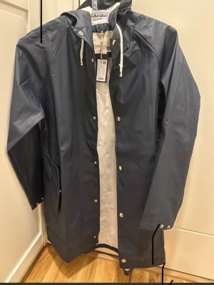 Mörkblå regnjacka från Lager 157 - Snygg mörkblå regnjacka med huva och vita snören från Lager 157. Jackan har långa ärmar, tryckknappar framtill och praktiska fickor. Perfekt för blöta dagar och enkel att matcha med din stil. Modellen heter Vänga Jill. 