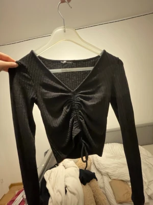 Svart ribbad topp med snörning - Säljer en svart långärmad topp från B Sister i storlek XS. Toppen har ribbad struktur, v-ringad hals och snygg snörning framtill som ger en rynkad effekt. Materialet är mjukt och stretchigt, perfekt för att skapa en cool look.
