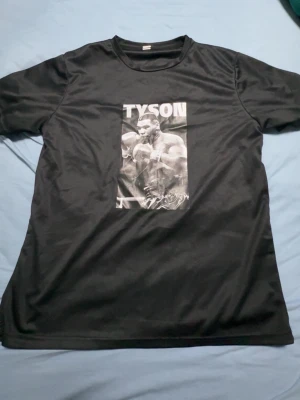 Svart t-shirt med Tyson-tryck - Cool svart t-shirt med ett stort tryck av boxaren Tyson på bröstet. T-shirten har rund halsringning och är kortärmad. Perfekt för dig som gillar sportiga och stilrena plagg med attityd.