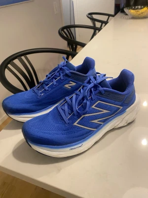 Blå New Balance Fresh Foam 1080 sneakers - Säljer ett par blå New Balance Fresh Foam 1080 sneakers med ventilerande mesh-överdel och vita, tjocka sulor. Skorna har snörning och stora silvriga N-loggor på sidorna. Insidan är neongul med Fresh Foam-tryck. Perfekta för träning eller promenader.