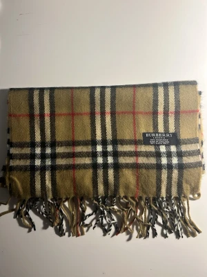 Rutig beige Burberry väst i kashmir - Säljer en klassisk rutig väst från Burberry i beige, svart, vitt och rött. Västen är tillverkad i 100% kashmir och har fransar längst ner. Den har det ikoniska Burberry-mönstret och är mjuk och lyxig, perfekt för att lyfta din outfit. (3A)