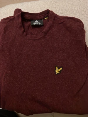 Vinröd stickad tröja från Lyle & Scott - Snygg vinröd stickad tröja från Lyle & Scott med klassisk rund hals och gul broderad logga på bröstet. Perfekt för dig som gillar stilrena och bekväma plagg. Tröjan har lång ärm och mjukt material som känns skönt mot huden.