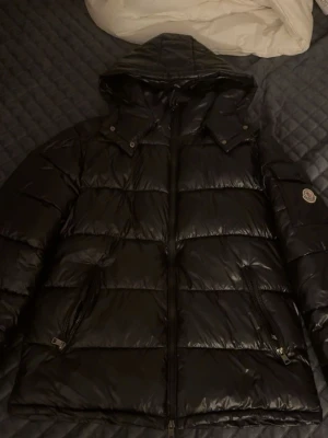 Svart pufferjacka från Moncler - Svart pufferjacka från Moncler med huva och dragkedja framtill. Jackan har två sidofickor med dragkedja och ett Moncler-märke på ärmen. Materialet är glansigt och känns lätt, perfekt för kalla dagar. Klassisk quiltad design och stilren look.
