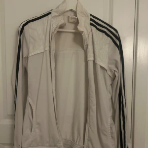 Vit Adidas vindjacka med svarta ränder - Säljer en vit vindjacka från Adidas i storlek S. Jackan har klassiska svarta ränder längs ärmarna, dragkedja framtill och två fickor. jackan har en sportig look med  långärmad design. Funkar bra som träningsplagg.