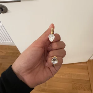 Guldfärgad armband med stora stenar - Säljer ett snyggt guldfärgat armband med tvinnad design och två stora, droppformade klara stenar i varje ände. Armbandet är öppet och flexibelt, perfekt för att ge lite extra bling till din outfit. Passar dig som gillar statement-smycken och glittriga detaljer.