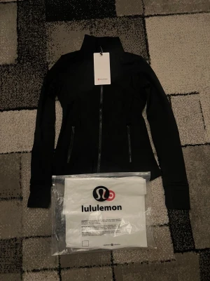 Svart träningsjacka från Lululemon - Snygg svart träningsjacka från Lululemon med hög krage och hel dragkedja framtill. Jackan har två praktiska fickor med dragkedja och är långärmad. Materialet är mjukt och stretchigt, perfekt för aktiva dagar. Stilren design med diskret logga på ryggen.