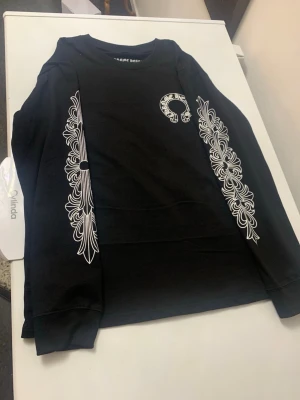 Svart långärmad t-shirt Chrome Hearts - Svart långärmad t-shirt från Chrome Hearts med vita grafiska tryck längs ärmarna och logga på bröstet. Stor Chrome Hearts-tryck på ryggen och CH-detalj vid nederkant. Skön bomullskvalitet och klassisk rund hals. Perfekt för dig som gillar streetwear med edge.