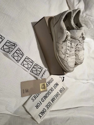 Golden Goose Ballstar - Golden Goose sneakers i vit färg med stjärndetalj på sidan. Klassisk låg modell med snörning och platt sula. Snyggt sliten look. Kommer med låda, dustbag, kvitto och etikett. Skorna är knappt använda och slitningarna är från fabrik. De vill säga att de är en del av designen som på många Golden Goose skor. Storlek 45. 