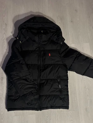 Svart Ralph Lauren puffer - Svart pufferjacka från Polo Ralph Lauren med huva, dragkedja och två sidofickor med zip. Jackan har det klassiska röda Polo-logot broderat på bröstet och är tillverkad i polyester. Perfekt för kalla dagar och har en schysst, modern look. Endast använd fåtal gånger och den är utan defekter.