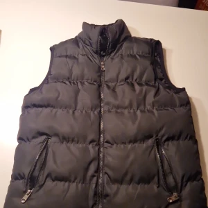 Svart dunväst med dragkedja - Säljer en svart dunväst med hög krage och två fickor med dragkedja framtill. Västen har quiltad design och stängs med en rejäl dragkedja i mitten. Perfekt att slänga över en hoodie eller tröja när det är kyligt ute.