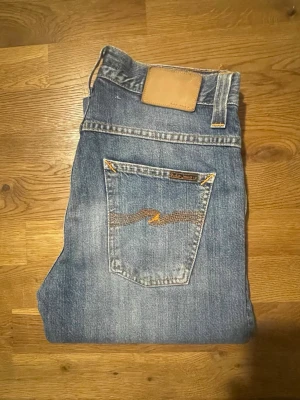 Blå jeans från Nudie med broderade fickor - Säljer ett par klassiska blå jeans från Nudie med snygga broderade detaljer på bakfickorna och en ljusbrun patch i midjan. Jeansen har rak passform och är tillverkade i slitstarkt bomullsmaterial. Perfekt för dig som gillar tidlös stil och schyssta detaljer.