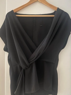 Svart omlottopp från Zara - Säljer en svart omlottopp från Zara i storlek S. Toppen har en djup v-ringning och knyt i midjan som ger en snygg siluett. Tillverkad i ett mjukt, lätt material som faller fint. Perfekt att styla med jeans eller kjol.