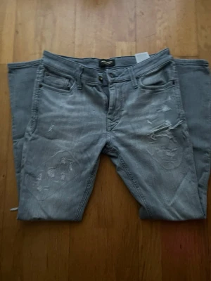 Grå jeans från Jack & Jones - Säljer ett par grå jeans från Jack & Jones med slitna detaljer och klassisk femficksdesign. Jeansen har en smal passform och är tillverkade i mjukt denimtyg med dragkedja och knapp framtill. Perfekt för dig som gillar en avslappnad men snygg stil. Dm mig för mer bilder, frågor eller prisförslag så löser vi nåt gött tsm