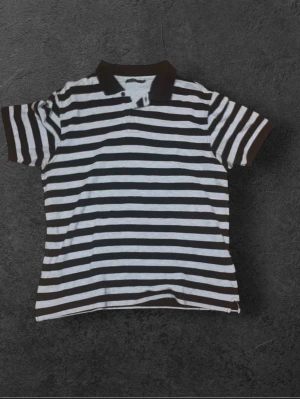 Randig svartvit pikétröja - Säljer en klassisk piké med breda svarta och vita ränder. T-shirten har korta ärmar, krage och knappslå framtill. Perfekt för dig som gillar en enkel men stilren look. Materialet känns som bomull och passformen är normal.
