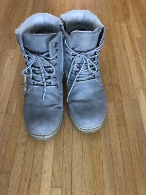 Grå fodrade kängor med snörning - Säljer ett par grå kängor med snörning och rund tå. Skorna har en mjuk, fodrad insida som håller fötterna varma och en platt sula för skön komfort. Utsidan är i skinnliknande material och de passar perfekt för kyliga dagar.