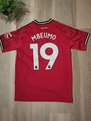 Manchester utd Mbeumo hemma - Manchester United hemma fotbollströja med Mbeumo och nummer 19 på ryggen, samt Adidas-logga och Snapdragon sponsor på framsidan. 