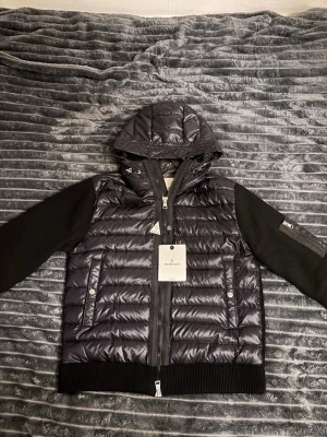 Moncler cardigan - Svart pufferjacka från Moncler med glansig yta, stickade svarta ärmar och huva med snörning. Jackan har dragkedja framtill, ribbade muddar och fickor med tryckknappar. Moncler-logga på ärmen och snygga detaljer i metall. L men sitter som M