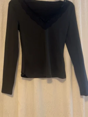 Svart långärmad topp med spets - Säljer en svart långärmad topp med v-ringad hals och dekorativ spetskant. Toppen har en slim passform och är tillverkad i mjukt material som känns skönt mot huden. Perfekt för dig som gillar stilrena plagg med en liten twist.