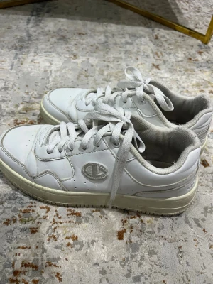 Vita sneakers från Champion - Stilrena vita sneakers från Champion, perfekta för vardagsbruk. Skorna är i bra skick och bekväma att använda. Det finns en mindre synlig missfärgning/ytmärke på sidan, vilket framgår av bilderna. Detta är endast kosmetiskt och påverkar inte funktion eller komfort. 