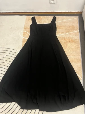 Svart långklänning med breda axelband - Säljer en elegant svart långklänning med breda axelband och utställd kjol. Klänningen har en klassisk och stilren design med markerad midja och fyrkantig urringning. Perfekt för dig som gillar tidlös stil och vill ha en klänning som känns både chic och bekväm.