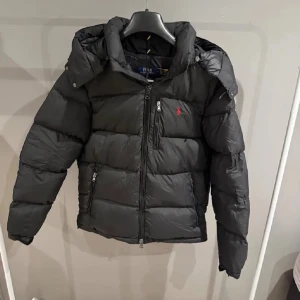 Svart Ralph Lauren jacka - Säljer en svart pufferjacka från Polo Ralph Lauren med huva och dragkedja framtill. Jackan har ett rött Polo-logo broderat på bröstet och flera praktiska fickor med dragkedja. Perfekt för kalla dagar och har en snygg, modern look.