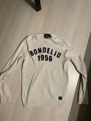 Vit tröja från Bondelid - Säljer en vit tröja från Bondelid med mörkblå text 'BONDELID 1956' på bröstet. Tröjan har rund halsringning, ribbade muddar och är långärmad. Perfekt för dig som gillar klassisk och enkel stil.