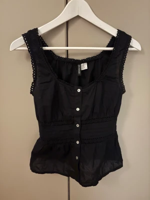 Svart blus från H&M Divided XXS - Säljer en svart blus från H&M Divided i storlek XXS. Blusen har knappar framtill, spetsdetaljer längs kanterna och midjan, samt en lätt rynkad och volangig nederdel. Ärmlös modell med rundad hals och dekorativ kant. Perfekt för sommarens alla tillfällen. Passar mig som vanligtvis har S