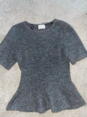 Grå stickad topp med peplum - Säljer en grå stickad topp från H&M med korta ärmar och peplum-detalj vid midjan. Mjuk och skön kvalitet som passar perfekt till jeans eller kjol. Rund halsringning och ribbad struktur ger en snygg och enkel look.