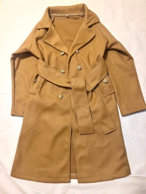Beige trenchcoat med bälte och knappar - Snygg beige trenchcoat med bred krage, dubbelknäppning med guldiga knappar och ett matchande bälte i midjan. Jackan har långa ärmar och två fickor framtill. Perfekt för dig som vill ha en klassisk och stilren look.