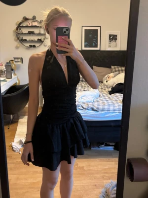 Svart kort halterneckklänning - Supersnygg svart kort klänning med halterneck och djup v-ringning. Klänningen har volangig kjol och öppen rygg, vilket ger en riktigt trendig och ungdomlig look. Perfekt för dig som vill sticka ut och känna dig extra fin.