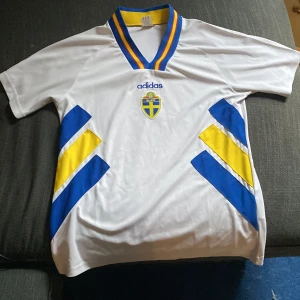 Sverige 94 retrotröja - Bilderna visar en vit Adidas fotbollströja med blå och gula detaljer, samt svensk landslagssköld. 