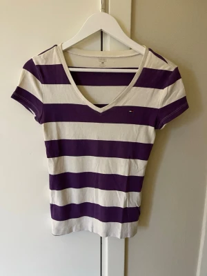 Randig t-shirt från Tommy Hilfiger - Säljer en snygg t-shirt från Tommy Hilfiger med breda lila och vita ränder. T-shirten har v-ringning, korta ärmar och en liten logga på bröstet. Materialet är mjuk bomull och passformen är slim. Perfekt för dig som gillar klassisk streetstil.