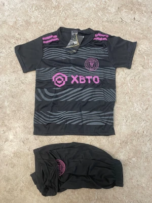 Svarta Inter Miami fotbollskläder Messi - Säljer ett svart Inter Miami fotbollskit med tröja och shorts. Tröjan har rosa detaljer, klubbmärke och sponsortryck framtill samt Messi och nummer 10 på ryggen. Shorts med klubbmärke i rosa. Materialet är polyester och designen har vågiga grå linjer.