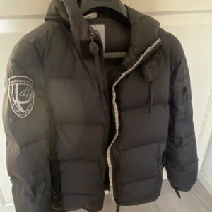 Svart dunjacka med huva - Snygg svart dunjacka med huva från Dbrand. Jackan har en dragkedja framtill och en  logga på ärmen. Perfekt nu till vintern. Andvänd fåtal gånger förra vintern!