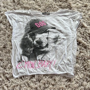 Vit t-shirt med hundtryck från Shana  - Vit t-shirt från Shana i bomull med korta ärmar. Stor svartvit print av en hund med keps där det står 'DOG' i rosa och texten 'C'MON BABY!!' i rosa nedtill. Avslappnad passform och rund halsringning. Perfekt för en cool och lekfull look. Perfekt till sommaren.  Tröjan är stretchig. Storlek XS