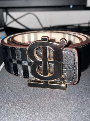 Svart rutig skinnväst med B-spänne - Snygg svart väst i skinn med rutigt mönster och ett stort metallspänne med B-logga framtill. Västen har detaljerade sömmar och insidan är beige med klassiskt rutmönster. Perfekt för dig som vill sticka ut med en lyxig accessoar.