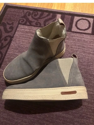 Grå mocka chelsea boots från Gant - Säljer ett par grå chelsea boots från Gant i mocka med beige elastiska paneler på sidorna och vit platt gummisula. Insidan är fodrad med mjukt material för extra komfort. Snygga dragflikar med Gant-logga upptill. Perfekt för kyliga dagar.