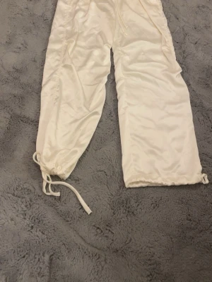 Vita cargo pants från VVR Couture - Snygga vita cargopants med glittriga detaljer på bakfickan och loggan i strass. Byxorna har dragsko vid bensluten så man kan antingen spänna åt dem eller ha dem lösa beroende på vad man tycker är snyggast och är tillverkade i ett glansigt syntetmaterial. Perfekt för dig som gillar statement-plagg och streetstyle. Kan eventuellt tänka mig gå ner i pris