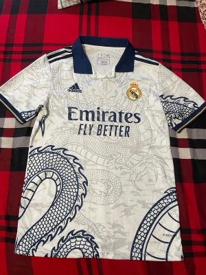 Real Madrid x Adidas draktröja S - Säljer en Real Madrid matchtröja från Adidas i slim fit, storlek S. Tröjan är vit med marinblå detaljer och har ett stort drakmönster över hela plagget. Klassisk krage och broderade loggor. Perfekt för dig som vill sticka ut på läktaren eller i stan.